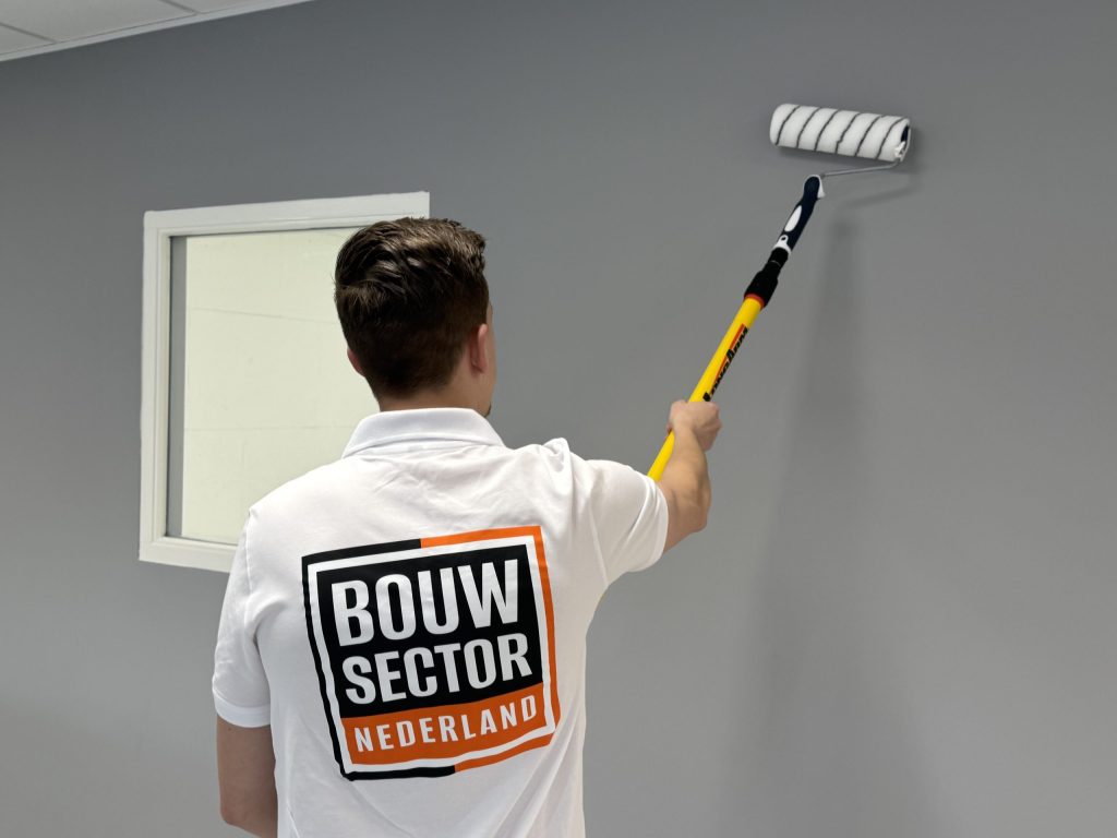 Schilder rolt grijze verf op een muur, links een raam in een wit geschilderd kozijn