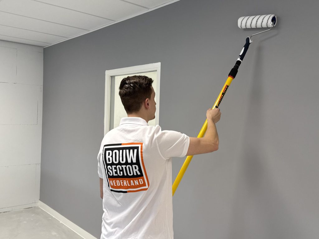 Schilder rolt met een verlengsteel grijze verf op een wand naast een wit binnenkozijn