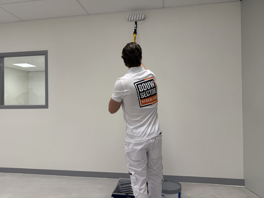 Schilder rolt de bovenrand van een lichte wand tot tegen het plafond, met verfbak en emmer op de vloer